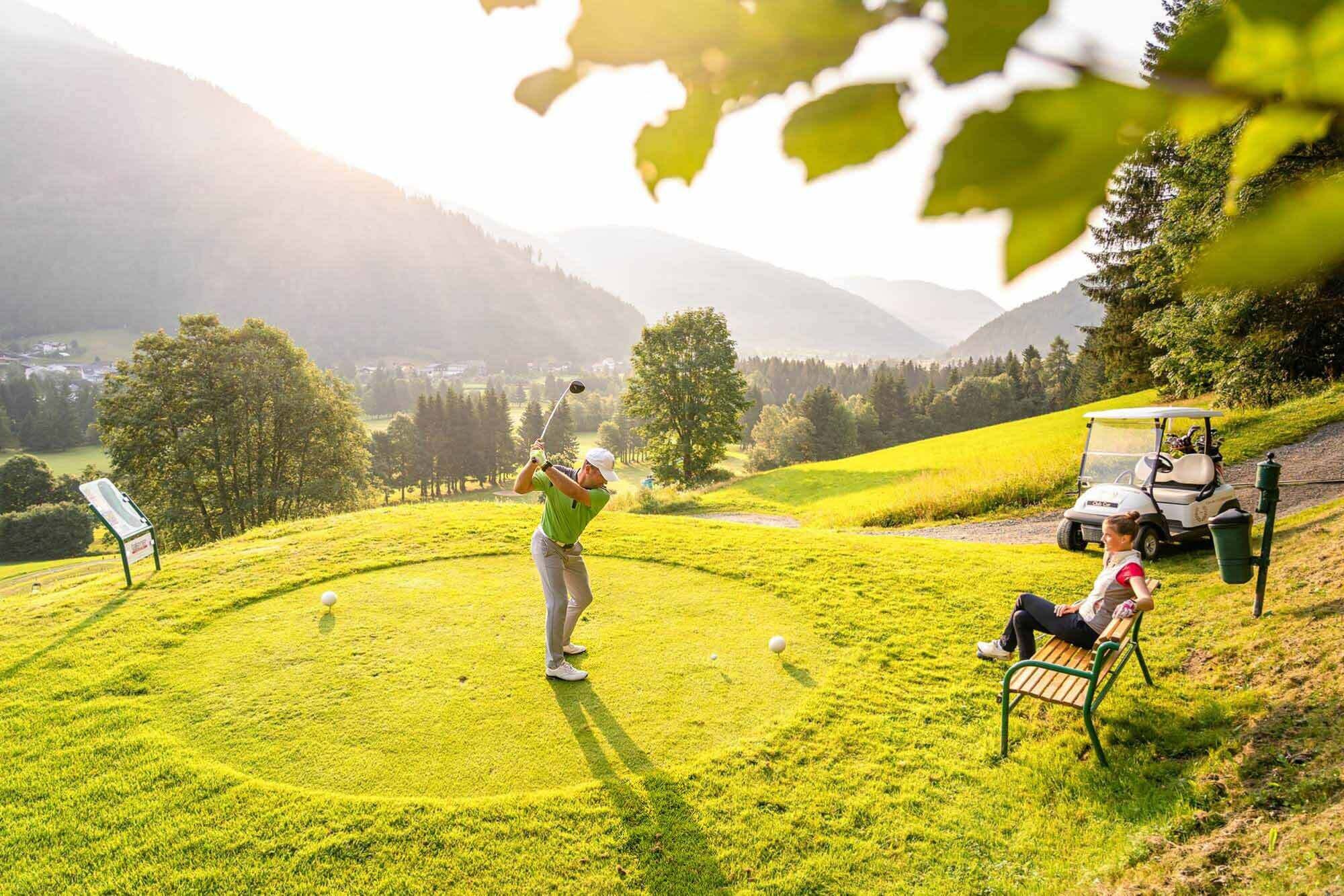 Eine Person die in der Golfarena in Bad Kleinkirchheim gerade zu einen Schlag ausholt und eine andere Person die auf einer Bank sitz und zu sieht.