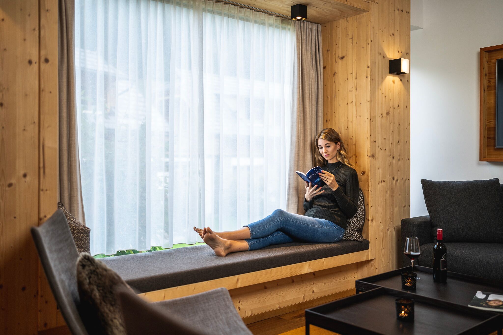 Eine Frau die im modernen Trattlerhof Chalet auf einen Sofa sitzt und ein Buch liest.