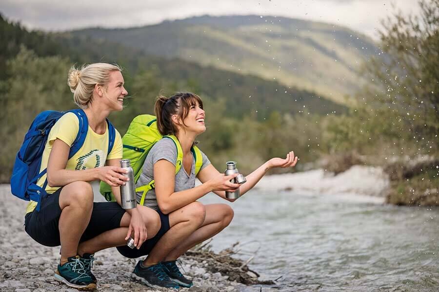 Zwei Frauen hocken an einem Fluss und sind gerade dabei, ihre Wasserflaschen mit Bachwasser zu füllen.