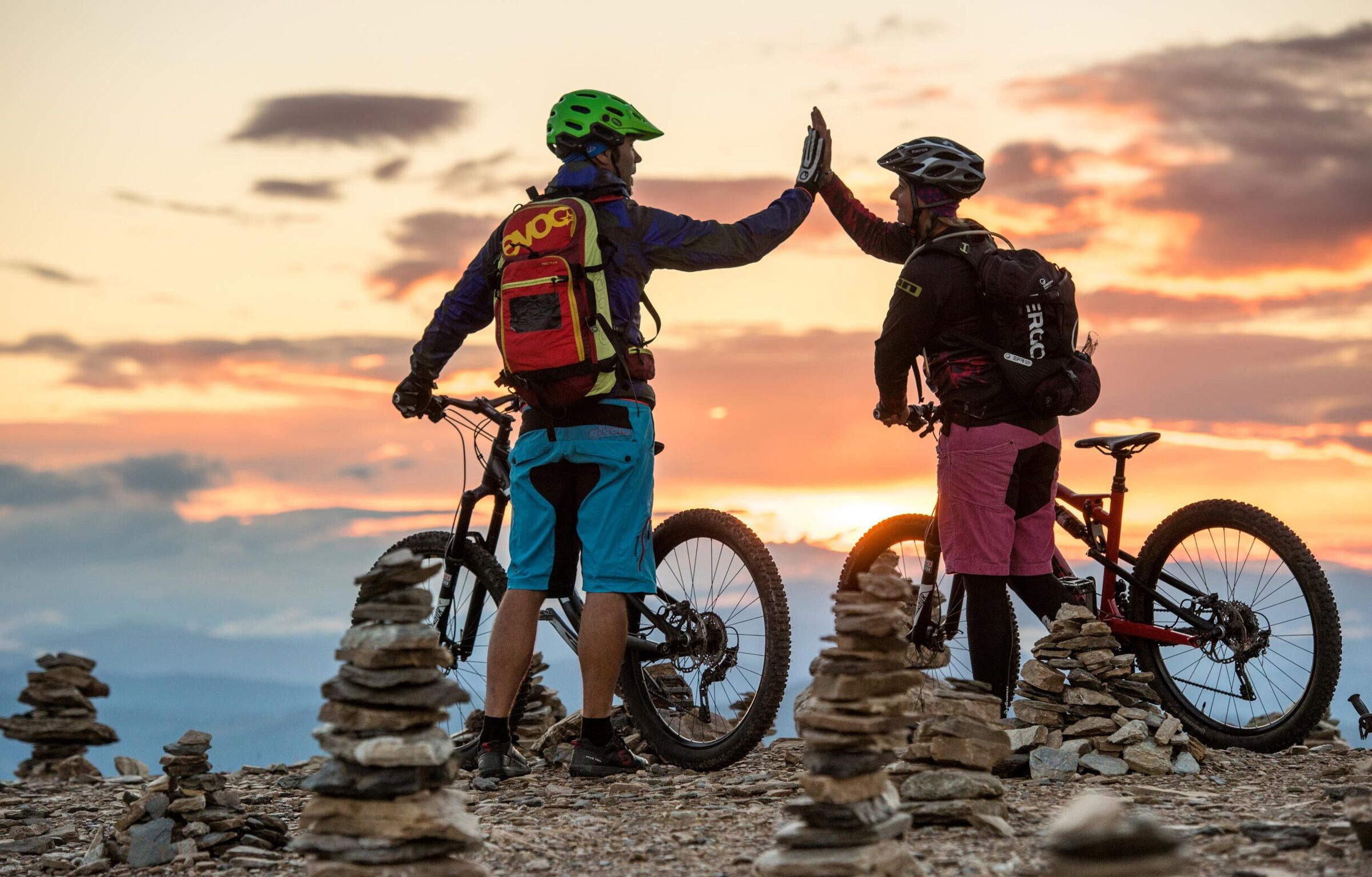 A couple is standing on a peak with their mountain bikes, high-fiving.