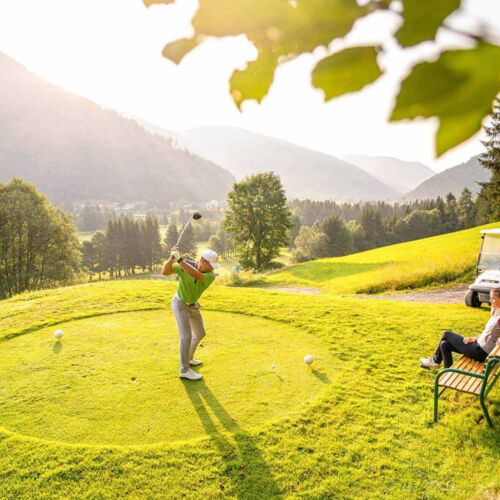 Una persona che fa uno swing nell'arena da golf di Bad Kleinkirchheim e un'altra persona seduta su una panchina che guarda.