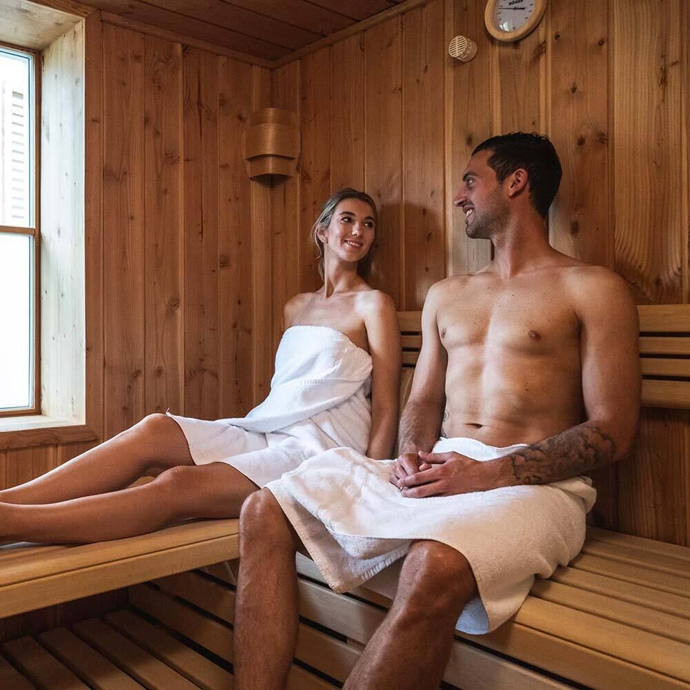 A woman and a man sit in the wooden sauna with panoramic view at Trattlers Hof-Chalets in Carinthia