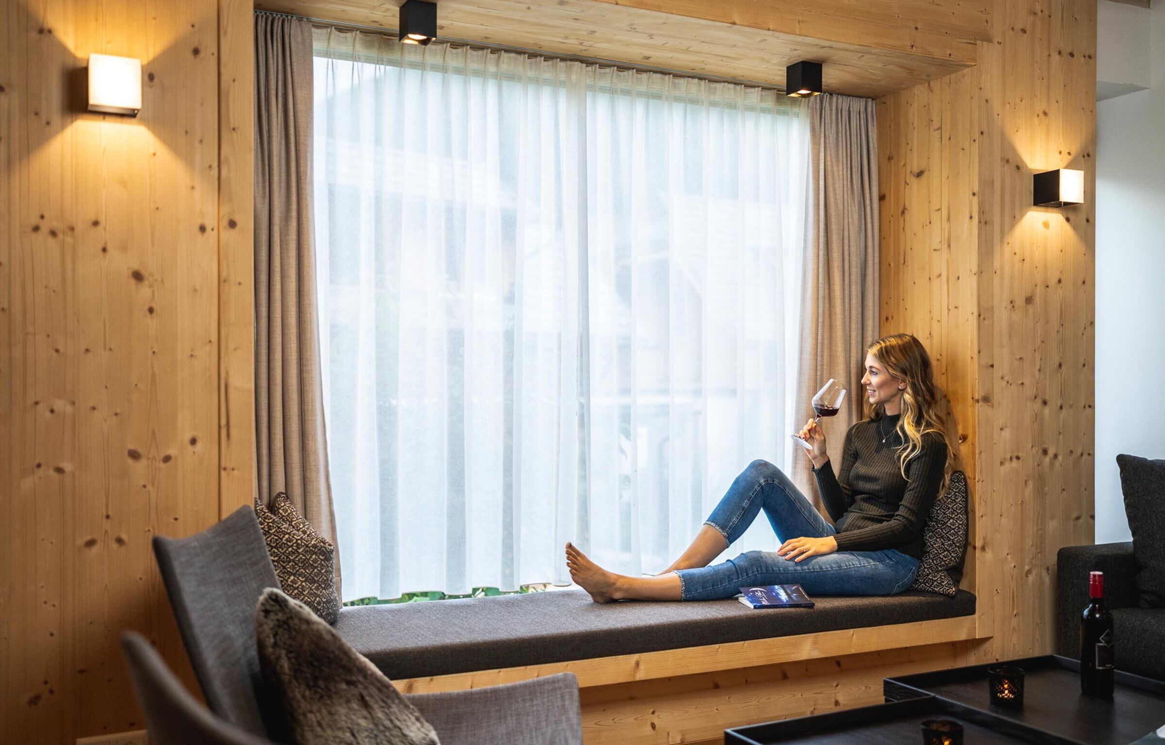 A woman sits in front of the panorama window in the chalet in Carinthia while drinking a wine