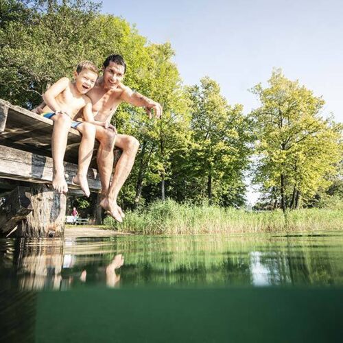 Ein Vater und sein Sohn sitzen am Steg und der Vater zeigt seinem Sohn etwas im Wasser des Millstätter Sees