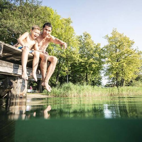 Un padre e suo figlio sono seduti sul molo e il padre sta mostrando al figlio qualcosa nell'acqua del lago Millstätter