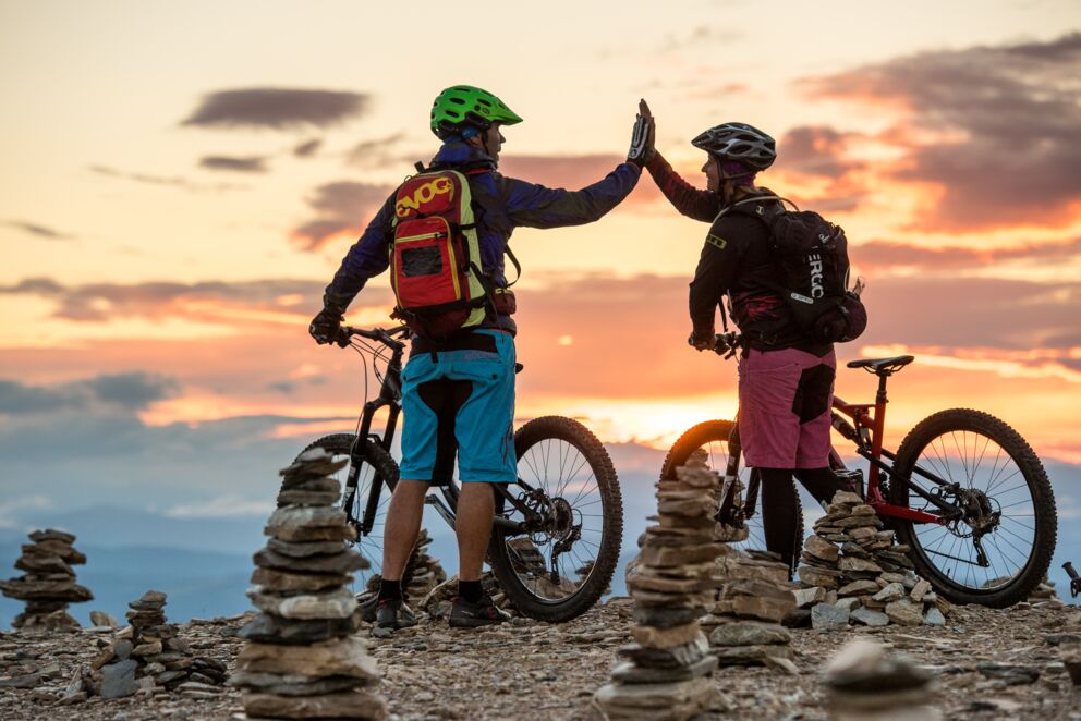 A couple is standing on a peak with their mountain bikes, high-fiving.