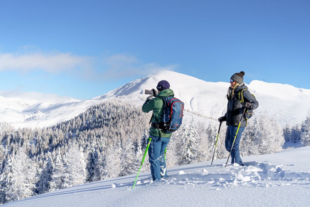 Due persone stanno ciaspolando nella neve alta e una persona sta fotografando con il cellulare le montagne innevate che si vedono.