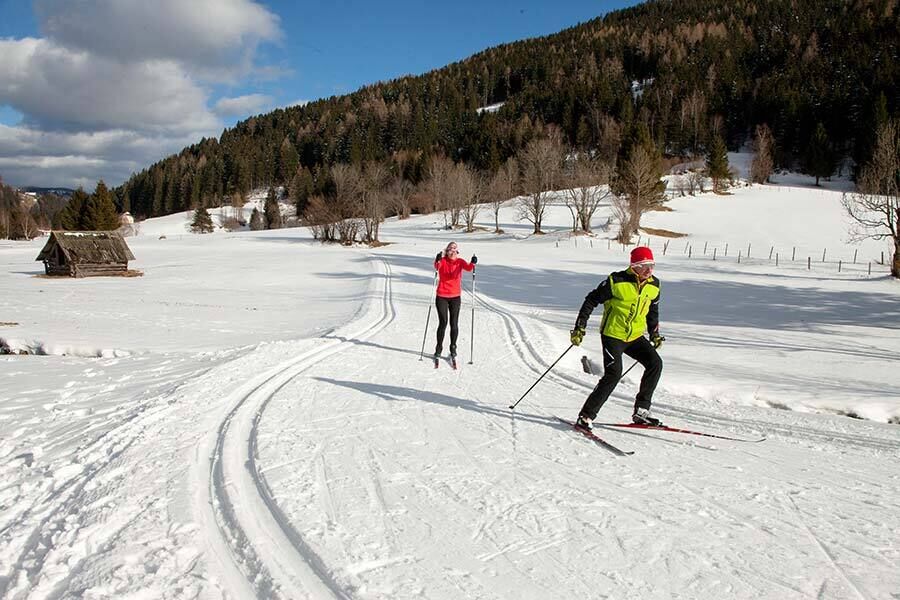Im Sonnenschein und auf präparierte Loipen in Kärnten gehen zwei Menschen den Wintersport Langlaufen nach