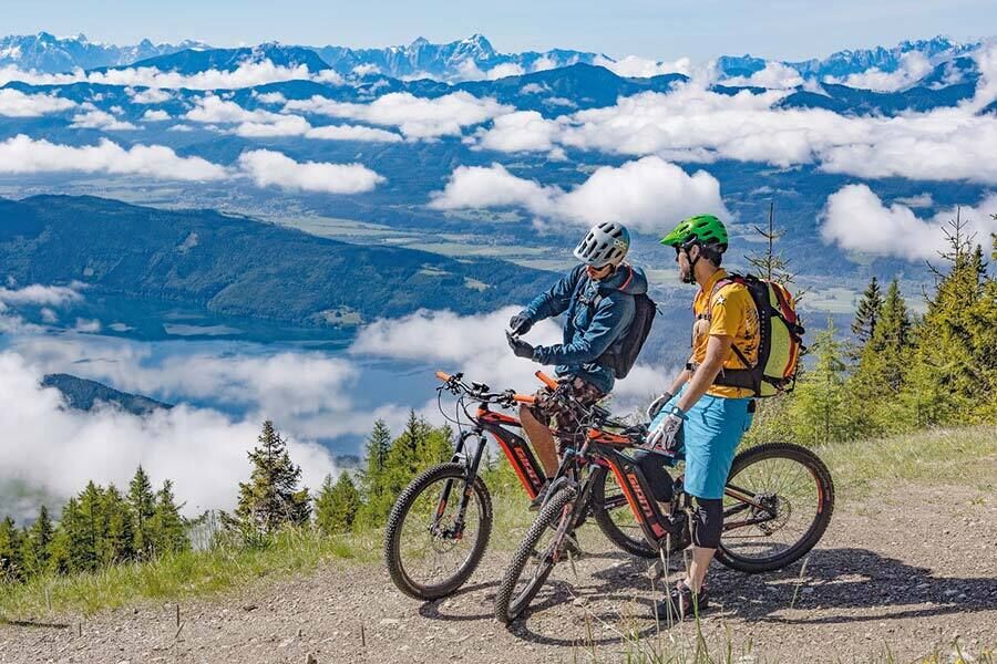 Two mountain bikers pause on the trail to discuss the route shown on their phone, with the valley and clouds stretching out behind them