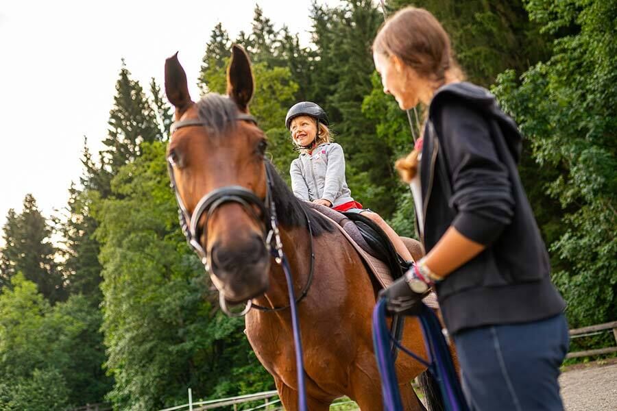 Ein kleines Mädchen das auf einen braunen gesattelten Pferd sitzt und die Reitlehrerin hält das Pferd am Strick.