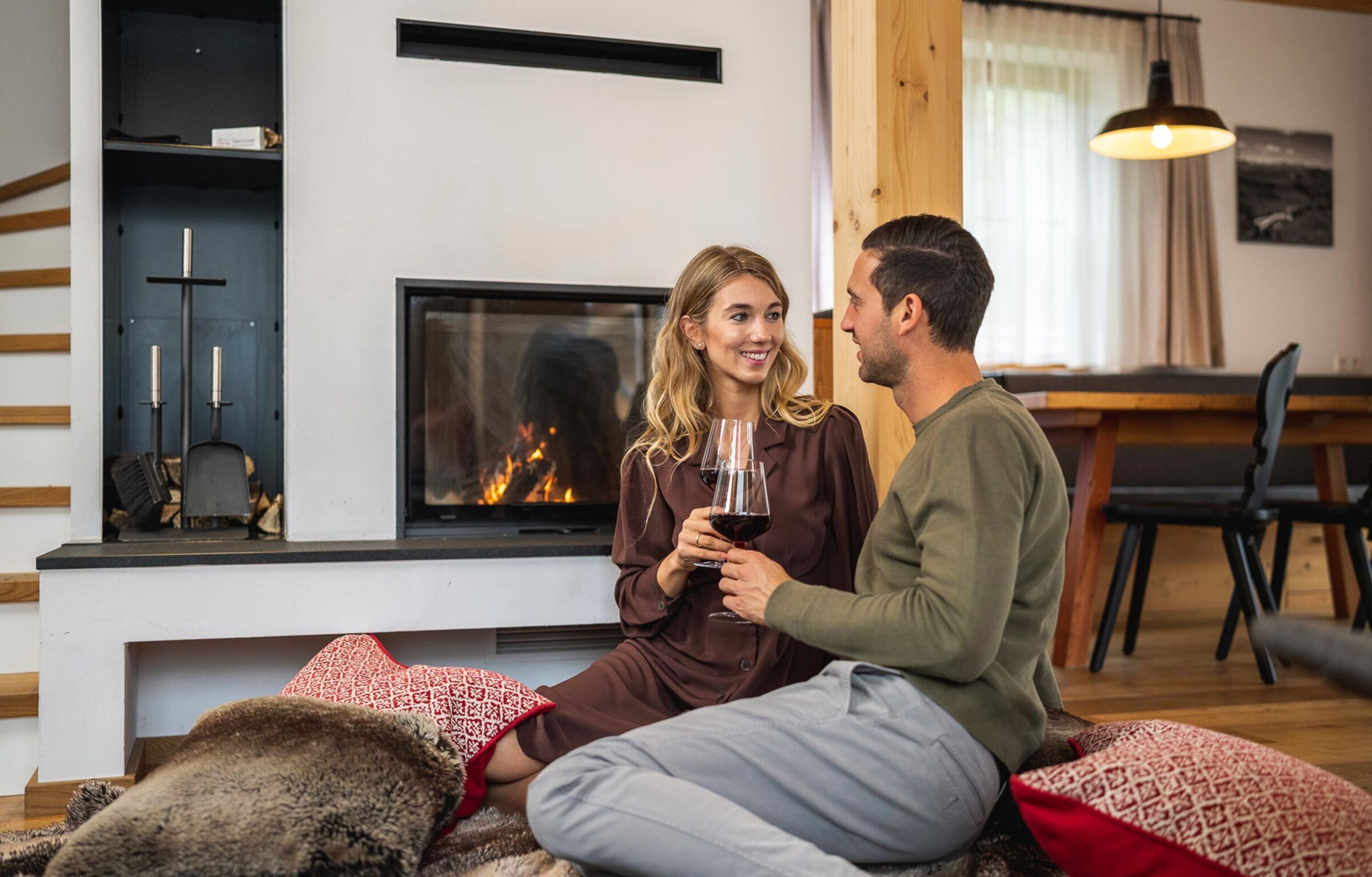 A couple smiling at each other and sitting in front of the fireplace with a glass of red wine