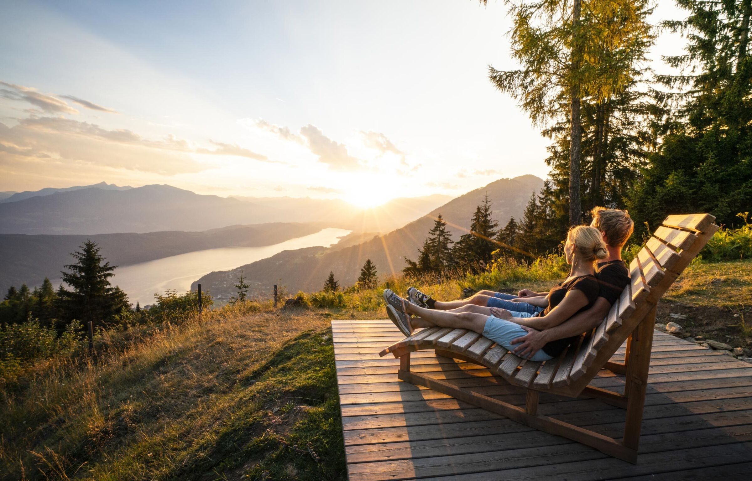 A couple sits on a wooden bench and enjoys the sunrise in the mountains.