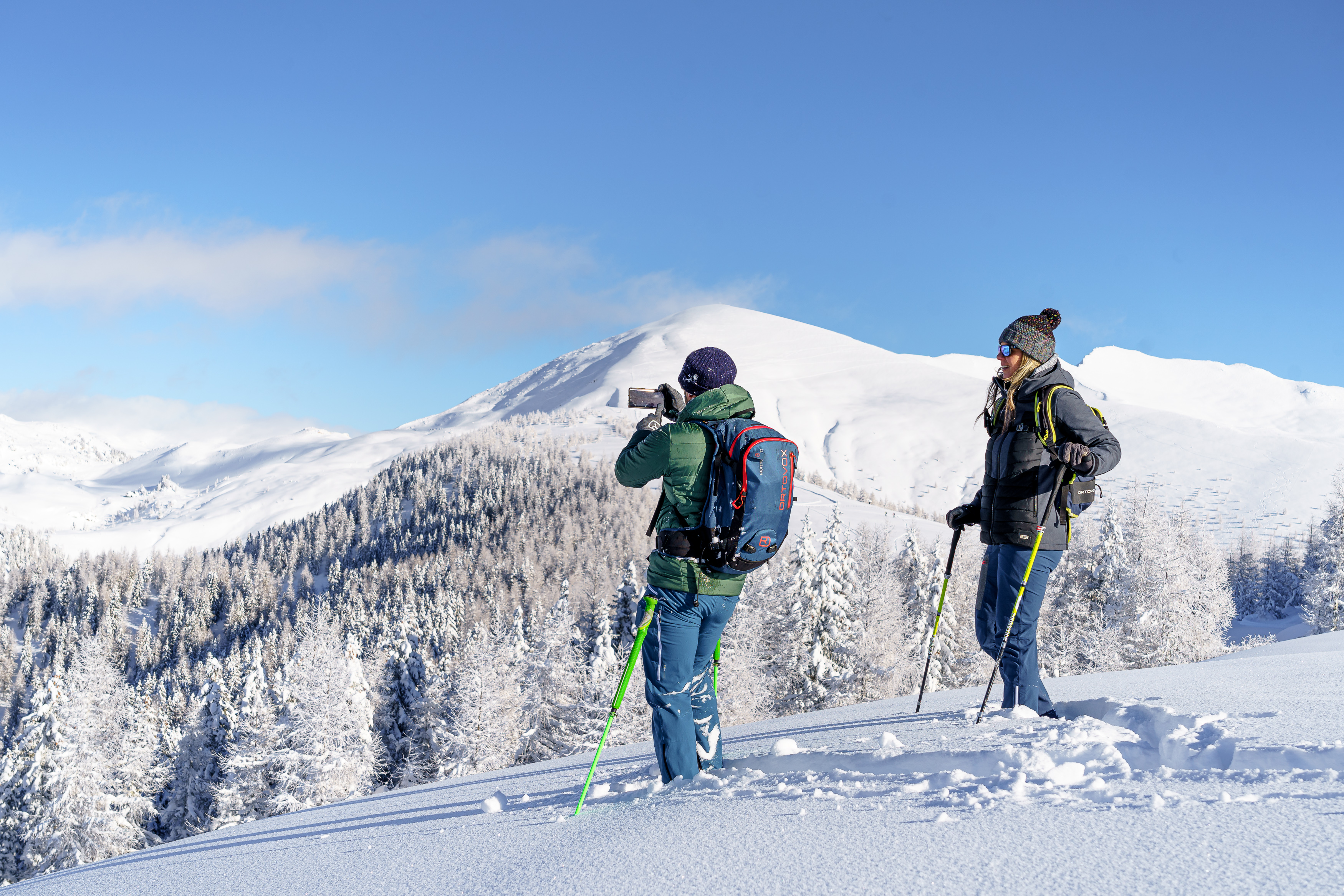 Two people are snowshoeing in deep snow and one person is taking a photo with the mobile phone of the snow-covered mountains that can be seen.