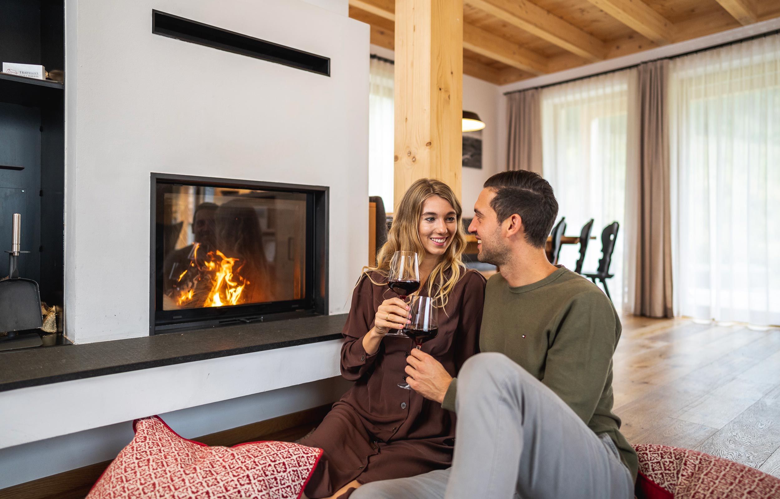 A couple is sitting in front of a fireplace drinking red wine.