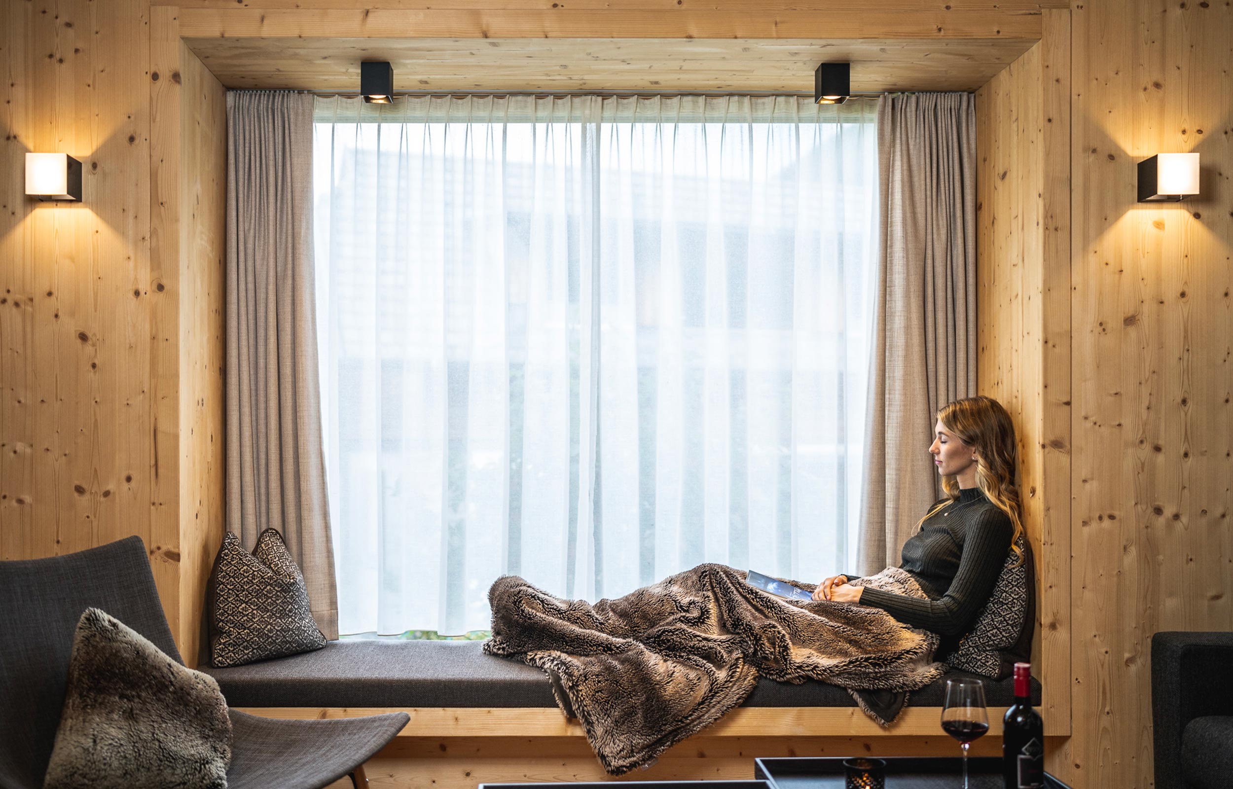Eine Frau die im Trattlerhof Chalet auf einen Sofa vorm Fenster sitzt und entspannt.